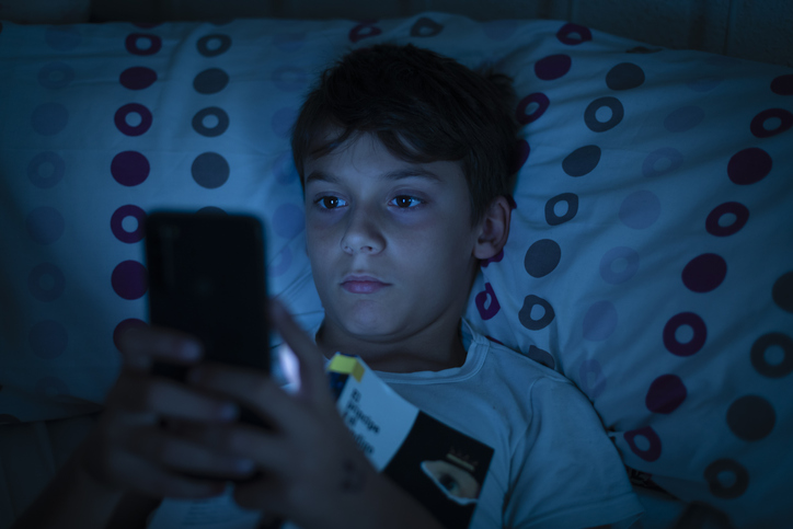 Child in bed at night with a book and a smartphone in the dark. Concept of problems with mobile addiction at young ages. Child with sleeping problems and insomnia due to the use of electronic devices
